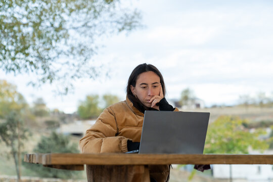 Young woman working on laptop at wooden table outdoors, focused and productive in casual fall attire, remote work lifestyle scene