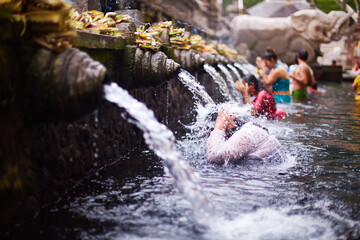 Bali, Indonesia, November 18, 2015. Woman participating in traditional water purification ritual at temple, kneeling under stone spout with water flowing over head, people in background praying