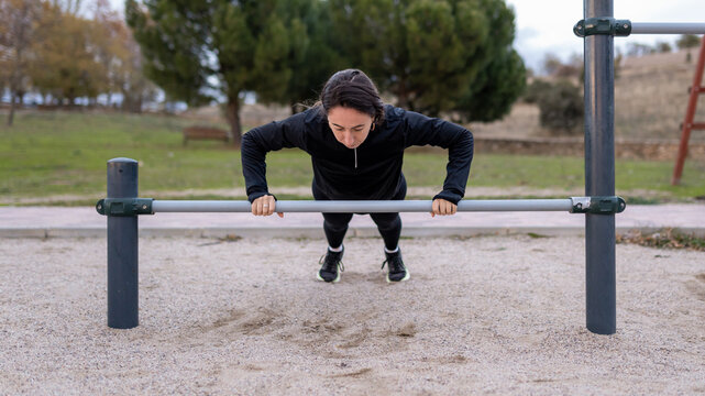 Woman performing outdoor calisthenics, engaging in strength training push-ups on parallel bars at a public park