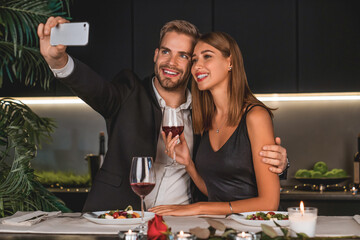 Happy couple having dinner and taking selfie using smartphone indoors. Vlogging and blogging for social media on a romantic date. Anniversary celebration.