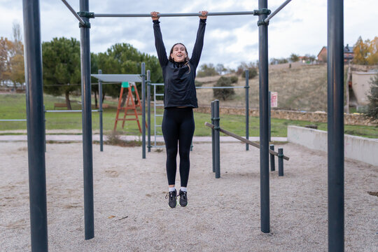 Young woman hanging from a pull-up bar, actively engaged in calisthenics training at an outdoor fitness park
