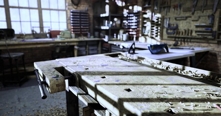 Dust and sunlight highlight a well used workbench in a woodworking shop. Tools line the walls, and a sense of craftsmanship fills the air, indicating a creative workspace.