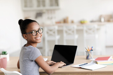 A smiling black girl wearing glasses sits at a table with a laptop at home, participating in an online meeting with her teacher. The atmosphere reflects social distancing during the pandemic.