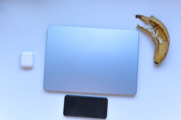 Minimal flat lay of a closed laptop, smartphone and wireless earbuds with a banana peel on a white background. Concept of digital detox, irony and lifestyle contrast.