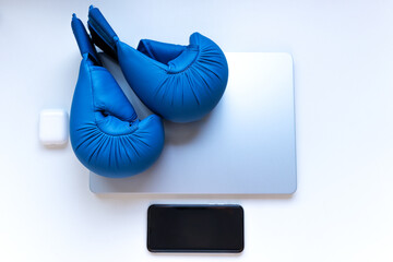 Blue boxing gloves placed on a closed laptop with smartphone and earbuds on a white surface. Concept of stress, work pressure, remote challenges and modern lifestyle.