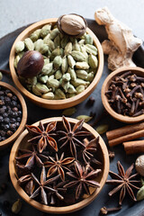 Close-up composition of ingredients for traditional Indian masala chai on black tray, featuring whole aromatic spices such as cardamom, cinnamon, cloves, black pepper, ginger on rustic background.