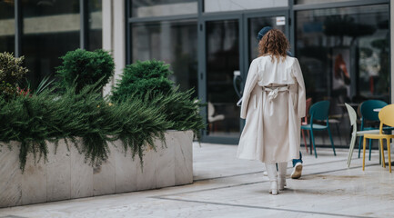 A stylish woman in a light beige trench exits past an outdoor cafe with colorful chairs and lush...