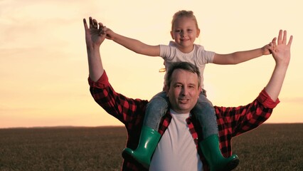 Smiling father and daughter walking at sunset agriculture wheat field closeup. Cute little girl sitting on male parent shoulders with open hands enjoy freedom happy childhood sunrise sky meadow