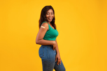 A vaccinated Black young woman poses confidently in a studio with a yellow background. She smiles and shows her arm with a medical plaster, promoting vaccination awareness and health safety.