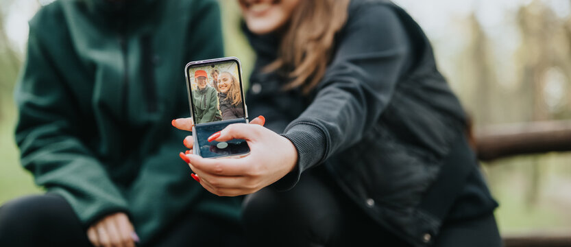 Two friends in jackets share a joyful moment outdoors, capturing a selfie on a smartphone. The park setting is blurred in the background, emphasizing the close, friendly vibe and casual lifestyle.