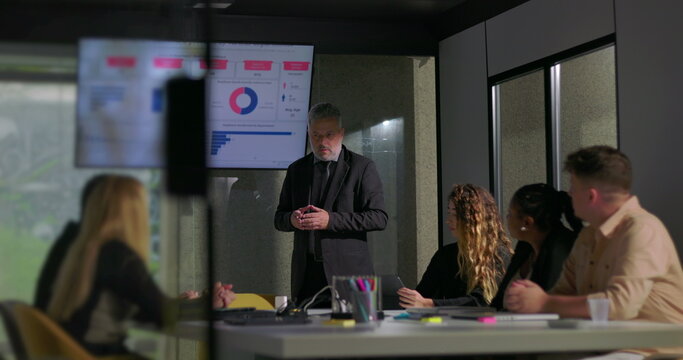 Corporate manager explaining report data to attentive colleagues in boardroom with screen displaying colorful charts and graphs