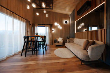 Cozy living dining area with vaulted timber walls, wide plank floors, beige sofa, large backlit mirror, orb pendants and sconces, round rug, accent chair, and tall black dining set.