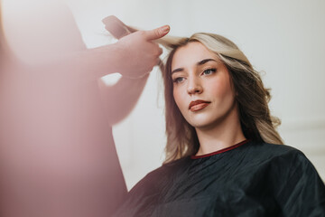 Naklejka premium A stylish woman sits in a hair studio wearing a cape as a stylist works on her hair, creating a polished, contemporary look. Warm lighting highlights her features in a calm, professional setting.