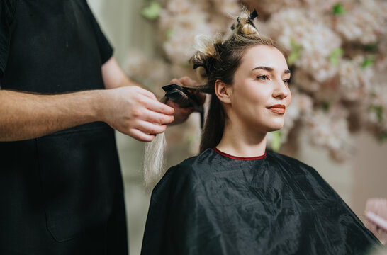 A woman in a black salon cape sits calmly as a stylist handles her hair, preparing a new style with extensions. Soft lighting and a floral backdrop create a chic, relaxing beauty session.