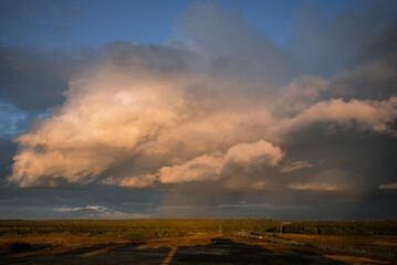 Golden sunlight painting dramatic storm clouds above a flat field and dark forest, creating a moody and powerful natural weather phenomenon during an evening sunset