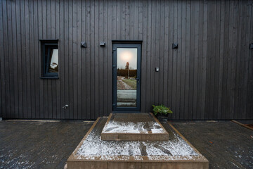 Black timber facade frames a narrow glass door, thin snow dusts tiered steps, small wall lights...