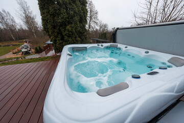 A bubbling hot tub churns turquoise water on a wet wooden deck, with leafless trees, hedges, and a playhouse. Low angle, wide lens, overcast light, shallow perspective.