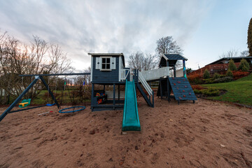 A DIY wooden playset with twin towers, raised walkway, teal slide, red grip wall, and swings sits on sand in a private yard at dusk in late autumn.