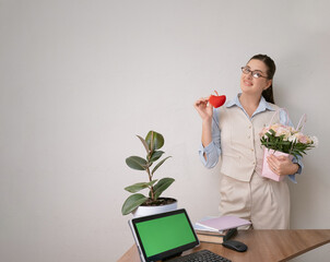 Surprised businesswoman in beige suit holding bouquet and tiny red heart, office workspace with green screen tablet