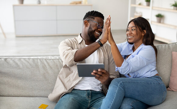 A cheerful black couple sits together on a couch in their living room. They are using a digital tablet and celebrating a success by giving each other a high five. Their joy is contagious.