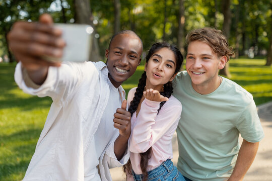 Three friends are having fun in a green park, smiling and posing for a selfie. They are capturing a joyful moment in bright daylight among trees.