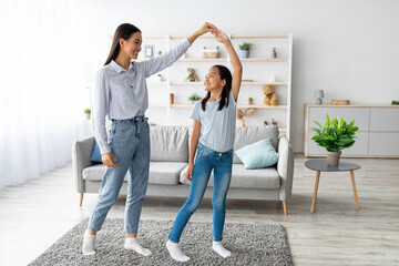 A cheerful mother and her daughter are dancing in their living room, holding hands and smiling....