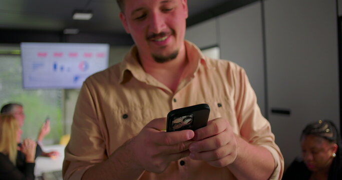 Close-up of young businessman smiling while reading message on smartphone, colleagues in background focused on laptops during meeting in office