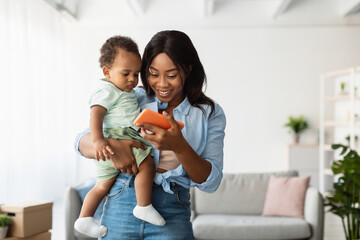 Young African American woman and her small son are smiling while looking at a mobile phone. The woman holds a baby as they relax together at home, engaging with fun and educational content.