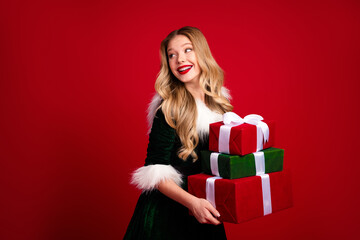 Festive girl in a velvet dress with white fur trim carries stacked red and green gifts against a red background for Christmas shopping