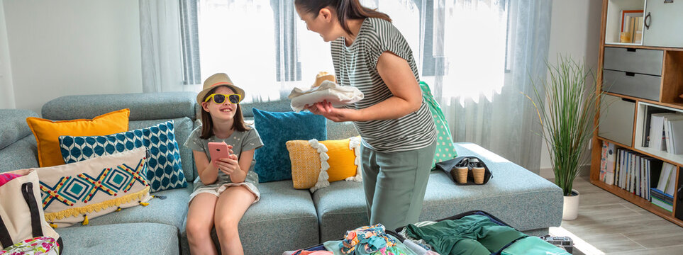 Banner of happy daughter with sunglasses and hat relaxes on sofa, using smartphone, while talking with mother that placing clothes into suitcase, preparing for family summer vacation in living room