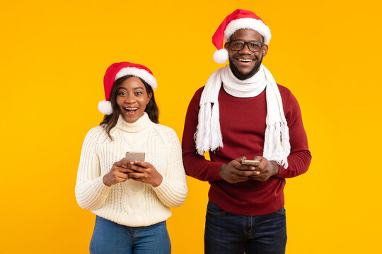 An African American couple joyfully holds smartphones in their hands while wearing festive Santa hats. They are posing in a studio against a yellow background during the Christmas season. - Powered by Adobe