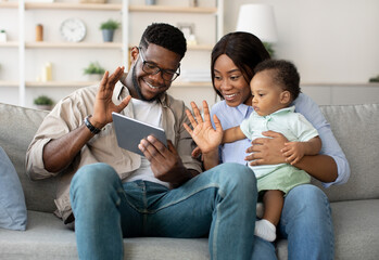 Cheerful African American family sits on a couch in their cozy living room. They are having a video call using a tablet, waving at the screen while the mother holds her baby boy.