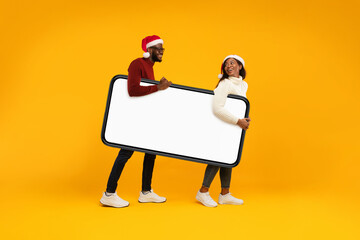 A joyful African couple wearing Santa hats stands against a bright yellow background, holding a large blank smartphone. They are preparing to promote Christmas offers creatively.