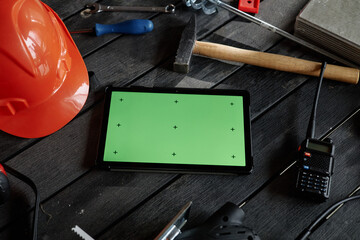 Tablet with green screen lying on wooden surface surrounded by construction tools including hammer, walkie talkie, hard hat, screwdriver, drill and metal ruler