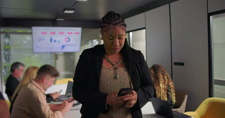 Concerned African American businesswoman looking at smartphone during office meeting, touching forehead while processing information, team in background
