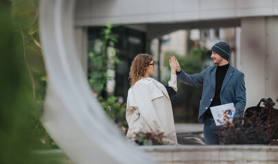 Two professionals share a high five on a city street, one holding a folder, dressed in smart casual attire. The scene conveys teamwork, collaboration, and positive business energy.