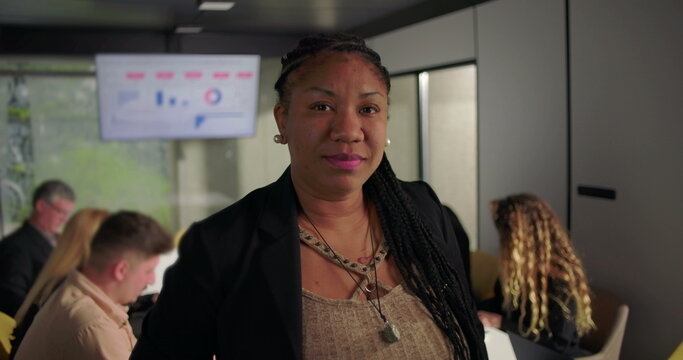 Confident African American businesswoman looking at camera in office, standing near conference table while team works in background on laptops and data charts