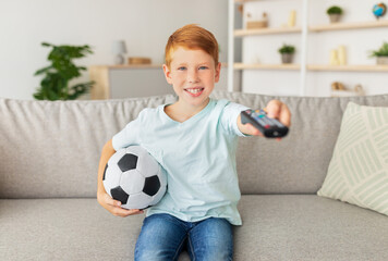 A cheerful redhead boy sits on a couch, holding a soccer ball and remote, as he watches his favorite football game at home. Excitement fills the room as he enjoys the match alone.