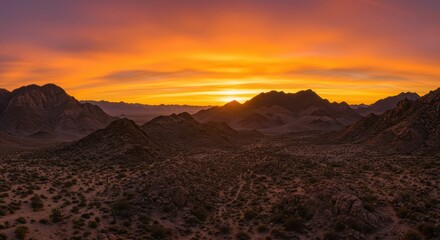 Naklejka premium Brilliant orange and yellow sunlight illuminates a vast, rugged desert mountain range at dusk.
