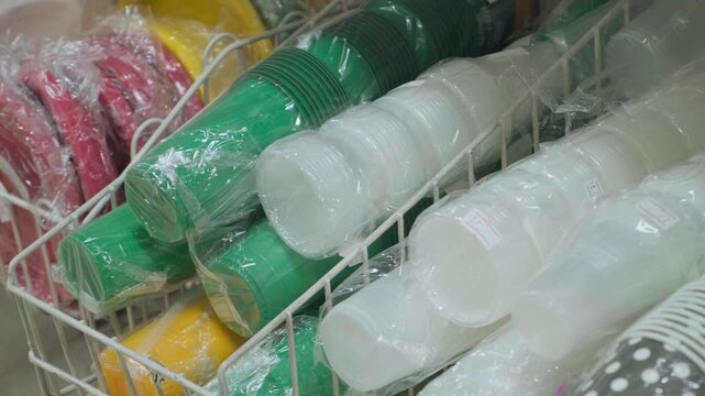 A close-up of various paper cups, plastic plates, colorful party supplies, and disposable tableware displayed on a grocery store shelf. Clutter, waste, environmental problems. Recycle plastic