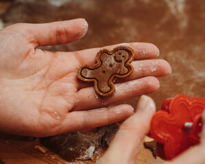 Close-up of a tiny gingerbread man cookie held in a flour-dusty hand. Detailed view of festive homemade Christmas baking, showing warm textures, cute shapes, and cozy holiday preparation.