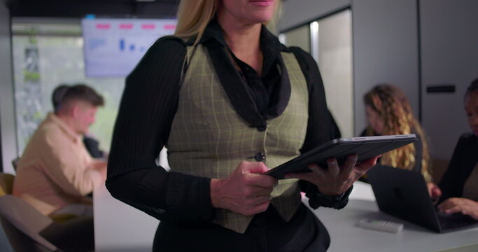 Confident businesswoman leading office meeting while reviewing information on tablet, supportive and focused team working in background, highlighting modern leadership style