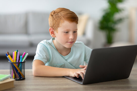 Ginger boy sits at a table in his home, focused on a laptop while completing homework. Stationery is nearby as he learns online during the COVID-19 pandemic.