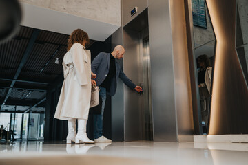 A man in a blue blazer and a woman in a white coat stand beside a polished elevator in a contemporary, glass-walled lobby, preparing to enter for a professional meeting.