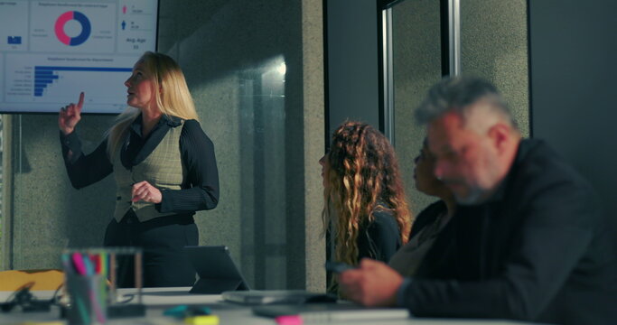 Confident businesswoman leading presentation during office meeting with team members listening, corporate environment with charts and data on display in modern workspace