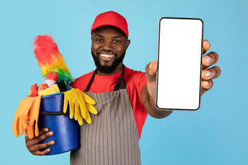 Black male cleaner holding basket with household tools and showing blank smartphone with white screen at camera, african american man recommending mobile app with cleaning service offers, mockup