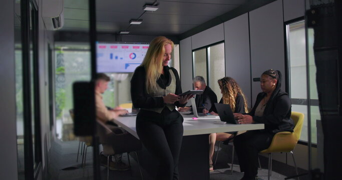 Confident businesswoman reviewing tablet during collaborative office meeting, team working in background with laptops, charts on screen, modern corporate environment - Powered by Adobe