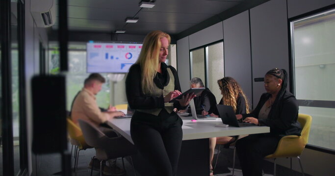 Confident businesswoman reviewing tablet during collaborative office meeting, team working in background with laptops, charts on screen, modern corporate environment