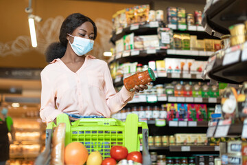 A woman inspects a jar of pasta sauce while shopping at a grocery store. She is wearing a mask and...