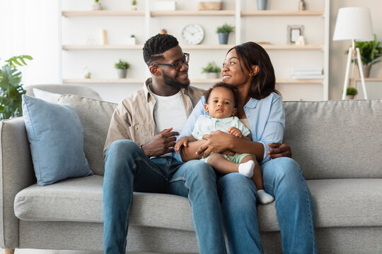 Loving millennial African American parents sit on a comfortable couch holding their adorable toddler son. They share joyful smiles, creating a warm family moment in their bright living room. - Powered by Adobe